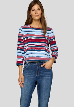 Frau trägt einen bunten gestreiften Langarmpullover und blaue Jeans, mit einer Hand in der Tasche, stehend vor einem hellgrauen Hintergrund.