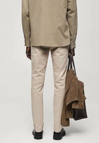 Pantalon beige ajusté, associé à une veste en daim marron. Le modèle tient un sac marron foncé et porte des chaussures noires.