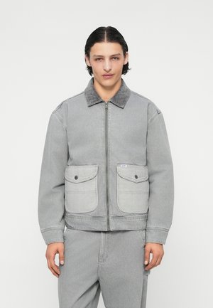 Veste en denim gris avec un col en velours côtelé, fermeture éclair sur le devant et deux grandes poches à rabat. Texture robuste, coupe décontractée et branding minimal.