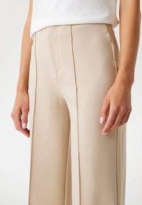 Pantalons larges beige en tissu lisse, dotés d'une poche avant et de coutures verticales pour plus de détails. Accentué par une coupe ajustée.