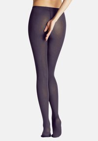 Modella che indossa collant trasparenti scuri, con una mano sul fianco, mostrando la vista posteriore dei collant su uno sfondo semplice.