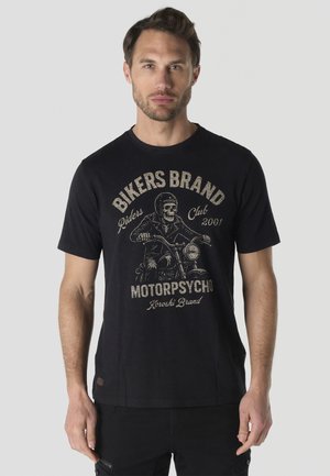 Hombre que lleva una camiseta negra con gráfico de jinete calavera y texto "Bikers Brand Riders Club 2001 Motorpsycho Koroshi Brand" de pie frente a un fondo liso.