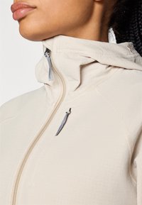 Veste zippée beige clair avec un col haut, texture lisse, comportant des détails de couture subtils et des tirettes de zip grises.