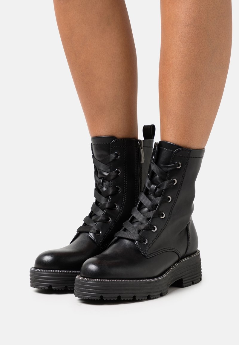 Personne portant des bottes de combat noires à lacets avec des semelles épaisses et des fermetures éclair latérales, debout sur un fond clair uni.