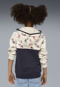 Sudadera con capucha que presenta la parte superior color crema con gráficos coloridos y una sección inferior de un oscuro tono navy. Los puños y el dobladillo son acanalados. Textura suave.