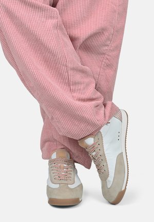 Gambe incrociate con pantaloni rosa a coste e scarpe da ginnastica bianche con dettagli in camoscio beige e lacci rosa decorati su uno sfondo chiaro.