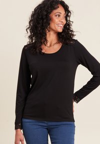 Schwarzes langärmliges Oberteil aus weichem Stoff, mit Spitzenbesatz an Schultern und Ärmeln, runder Ausschnitt, kombiniert mit blauen Jeans.