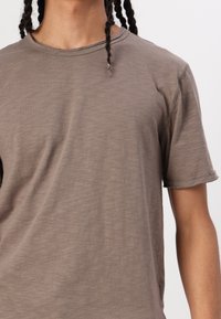 Braunes, kurzärmliges T-Shirt aus leichtem Stoff mit Rundhalsausschnitt und rohen Saumdetails; weiche Textur mit leichtem Glanz.