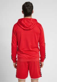 Haut de sport rouge avec une surface texturée, muni d'une capuche et d'accents noirs à motifs sur les manches, associé à un short rouge.
