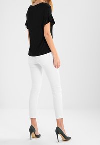 Žena stojící, oblečená v černém topu s krátkým rukávem, bílých slim-fit kalhotách a černých vysokých podpatcích na jednobarevném pozadí.
