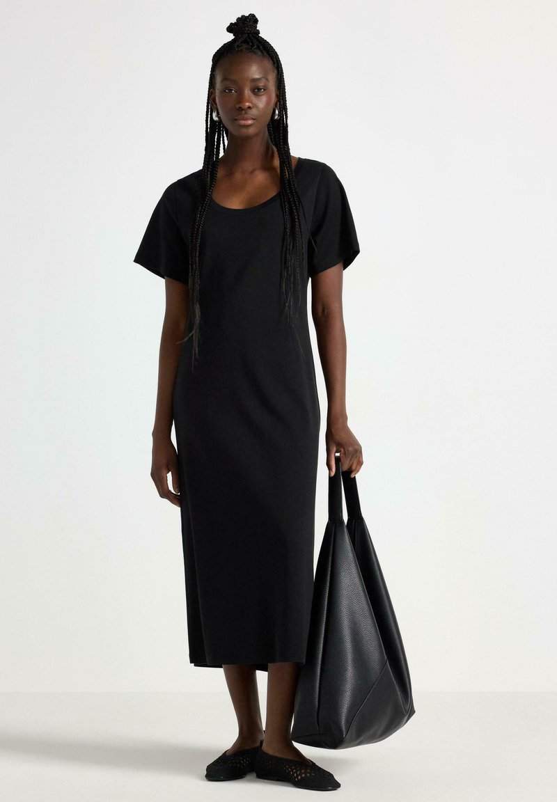 Abito maxi in cotone nero con maniche corte, scollo a barchetta e vestibilità comoda. La modella tiene una grande borsa a tracolla in pelle nera.