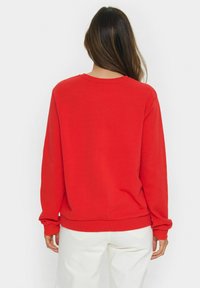 Rotes Sweatshirt mit langen Ärmeln, Rundhalsausschnitt und geripptem Saum. Weicher Stoff, Rückansicht, kombiniert mit weißen Hosen. Einfaches Design, keine Muster.