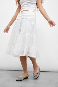 Woman wearing white tiered skirt and sleeveless top, standing with colorful floral embroidered slip-on shoes visible.