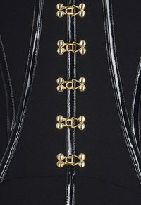 Schwarzes Kleidungsstück mit glänzenden schwarzen Kunstlederdetails und goldfarbenen Hardwareelementen, die im vertikalen Abstand entlang der Mitte verlaufen.