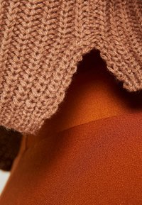 Pull en maille marron chaud avec un motif texturé, superposé sur un tissu lisse de couleur rouille, mettant en valeur les matériaux contrastés.