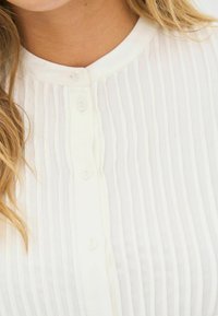 Hvid plisseret bluse med rund halsudskæring, der har fem knaplukninger og lodrette teksturdetailler, lavet af letvægtsstof.