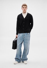 Cardigã preto com decote em V e botões, combinado com jeans soltos azul claro. Segurando uma pequena bolsa preta. Ténis brancos nos pés.