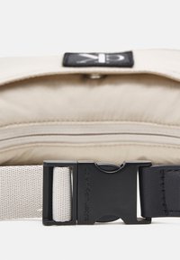 Bolsa de cintura de tela beige con hebilla de plástico negra. Presenta una textura suave, costuras lineales y una etiqueta con el logo en la parte superior.