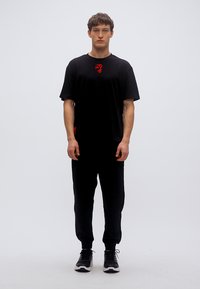 Μαύρο μπλουζάκι με κόκκινο λογότυπο, σε συνδυασμό με μαύρο παντελόνι φόρμας και μαύρα αθλητικά παπούτσια. Λείο ύφασμα, άνετη εφαρμογή και μίνιμαλ σχέδιο.