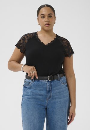 Kobieta w czarnej bluzce z koronkowymi rękawami, włożonej do niebieskich, wysokich dżinsów z czarnym paskiem, stojąca na tle jasnej, jednolitej ściany.