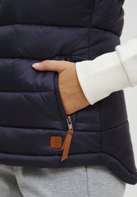 Námořnická prošívaná vesta s kapsou na zip, s koženou akcentní etiketou, hladkým povrchem a prošívaným designem. Pod ní bílá košile s dlouhým rukávem.