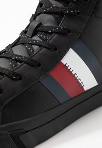 Zwarte high-top sneaker van glad leer. Heeft zwarte veters, een witte en rode streep met de tekst 'HILFIGER'. Robuuste rubberen zool.