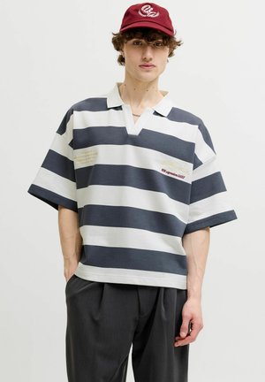 Persoană tânără purtând o șapcă roșie, un tricou polo supradimensionat cu mânecă scurtă cu dungi orizontale bleumarin și albe și pantaloni largi gri închis.