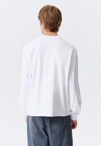 Person with short light brown hair wearing a loose white long-sleeve shirt and blue jeans, viewed from behind against a plain white background.