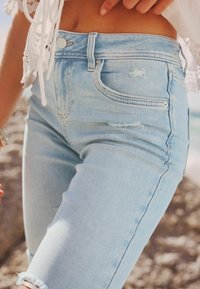 Vaqueros de mezclilla azul claro con una cintura alta, que presentan una textura desgastada, bolsillos frontales y dobladillos deshilachados. Detalles de hardware mínimos.