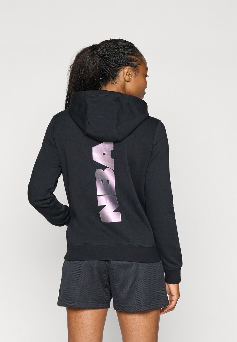 Svart hettegenser med metallisk rosa "NBA"-design på baksiden. Har lange ermer og et teksturert stoff. Kombinert med svarte shorts.