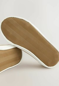 La vista de dos zapatillas posicionadas al revés, mostrando sus suelas de goma beige con un patrón de espiga sobre un fondo claro.