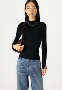 Črna rebrasta dolgosleeve majica z visokim ovratnikom in stranskimi razpoki, v kombinaciji s svetlo modrimi strganimi kavbojkami in temno rjavim torbico za ramo.