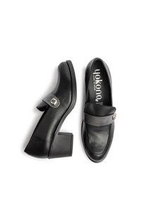 Mocasines de tacón de cuero negro con punta redonda, una correa ancha en el empeine y un detalle de hebilla metálica circular. Tacón de bloque texturizado.