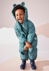 Combinaison de neige matelassée turquoise avec oreilles d'ours, dotée d'une fermeture éclair, de poignets élastiques et de bottes en caoutchouc navy. L'arrière-plan présente des couleurs douces et neutres.