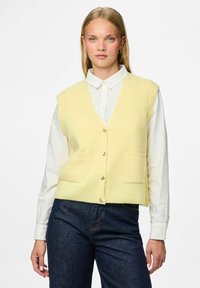 Žuta rebrasta pletena prsluk s dubokim V-izrezom, prednjim kopčanjem na dugmad i dva našivena džepa, nosi se preko bijele košulje s kragnom.