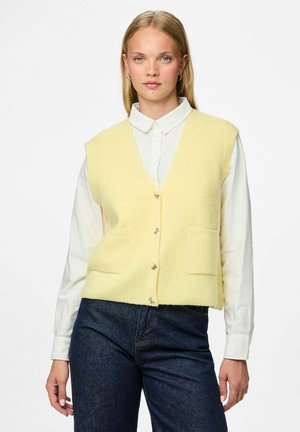 Gilet en tricot côtelé jaune avec un décolleté en V profond, fermeture à boutons sur le devant et deux poches plaquées, porté par-dessus une chemise blanche à col.
