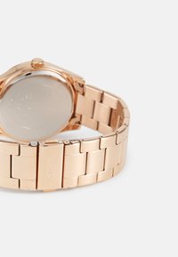Montre en métal rose gold avec un bracelet à maillons, dotée d'une finition polie, d'un boîtier rond et d'une fermeture à clip. Des inscriptions au dos sont visibles.