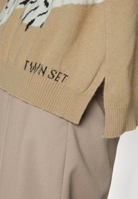 Pull beige en maille avec un motif blanc et noir en haut, présentant un ourlet côtelé et des fentes sur les côtés. Marque "TWIN SET" inscrite en bas.
