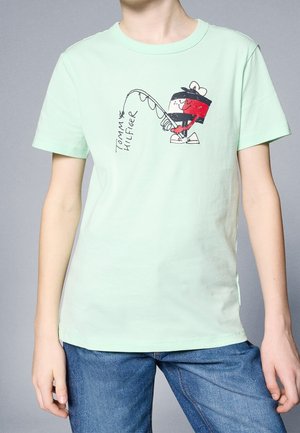 Mátově zelené tričko s krátkým rukávem s kreslenou postavičkou, která rybaří, s černými, červenými a bílými detaily. Text zní "Tommy Hilfiger."