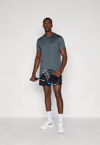 Mann står og holder en tennisracket iført grå skjorte, svarte shorts, hvite sokker og hvite tennissko foran en ensfarget bakgrunn.