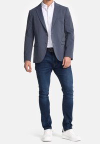 Grauer Blazer mit zwei Knöpfen, Klappentaschen, getragen über einem weißen Hemd und dunklen Jeans, kombiniert mit weißen Sneakern. Glatte Stoffstruktur.