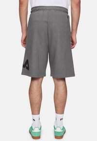 Pantalones cortos de algodón gris con una cinturilla elástica. Presenta un logo negro en el lado izquierdo. Se lleva con zapatillas verdes y calcetines blancos.