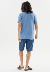 Vaaleansininen polo-paita yhdistettynä rullattuihin tummansinisiin shortseihin ja ruskeisiin liukuviin sandaaleihin. Sulava kangas ja rento malli.