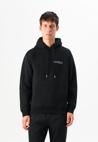 Young man stands wearing a black hoodie with white text "Replay Denim Co. Since 1981" on chest, black pants, neutral expression, white background.