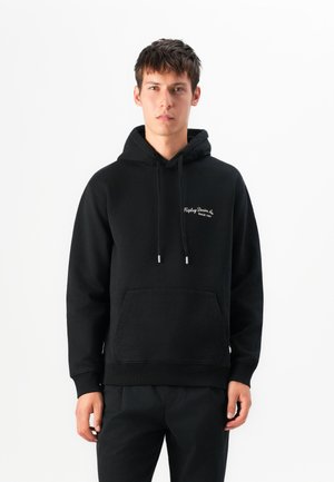 Young man stands wearing a black hoodie with white text "Replay Denim Co. Since 1981" on chest, black pants, neutral expression, white background.