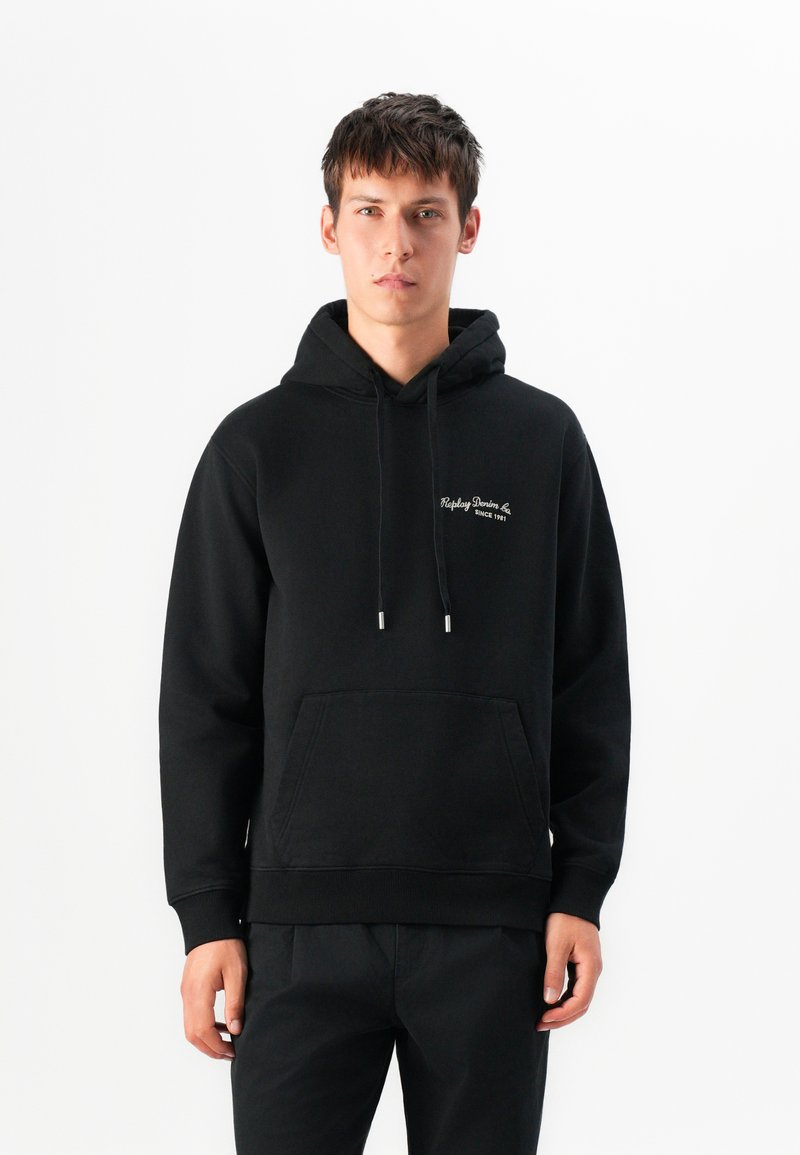 Young man stands wearing a black hoodie with white text "Replay Denim Co. Since 1981" on chest, black pants, neutral expression, white background.