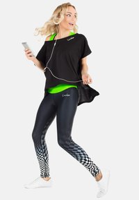 O bluză oversized neagră peste un top verde, colanți negri cu un model în carouri și adidași albi. Persoana ține un telefon, purtând căști.