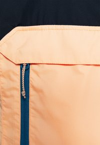 Poche zippée sur un tissu orange à finition texturée, bordée de matière bleu marine. Comprend une fermeture éclair bleue contrastante avec un curseur.