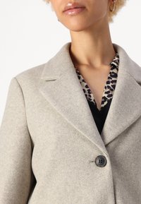 Cappotto grigio chiaro in lana con colletto a scalfo e chiusura a un bottone. Indossato sopra una camicia con stampa leopardata e scollo a V nera.