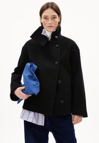 Schwarzer Wollmantel mit hohem Kragen und sechs schwarzen Knöpfen. Das Model hält eine blaue Tasche. Darunter sind ein gestreiftes Hemd und eine Cordhose sichtbar.